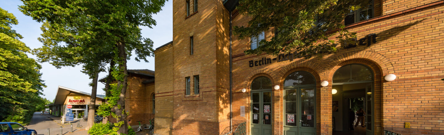 Das Bild zeigt ein Gebäude aus gelblichen Ziegelsteinen mit der Aufschrift "Berlin-Dahlem" über dem Eingang. Der Eingang hat drei große, grün gestrichene Doppeltüren mit runden Leuchten. Links neben dem Gebäude ist ein Zweig einer Straße zu sehen, an der ein Alnatura Super Natur Markt liegt. Große, grüne Bäume säumen die Straße und spenden Schatten.