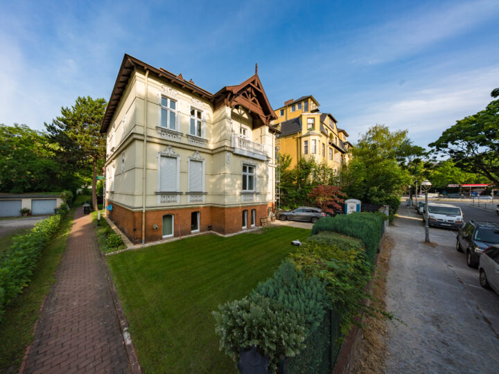 Das Bild zeigt eine charmante, mehrstöckige Villa mit gelber Fassade und roten Ziegelsteinen im Erdgeschoss. Umgeben von grüner Vegetation und einem gepflegten Garten liegt das Gebäude in einer ruhigen Wohnstraße. Im Hintergrund sind weitere historische Gebäude sichtbar. Der Himmel ist blau mit wenigen Wolken.