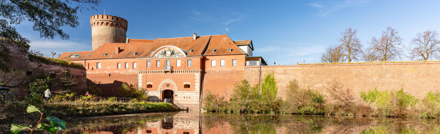 Historische rote Festungsanlage mit rundem Turm, hohen Backsteinmauern und Wassergraben, dahinter Bäume unter blauem Himmel.