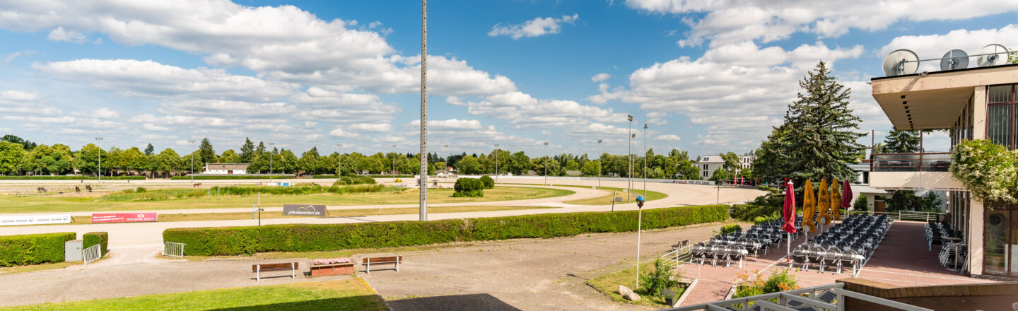 Ein sonniger Tag auf der Trabrennbahn Mariendorf. Im Vordergrund ein Bereich mit gestapelten Stühlen und Sonnenschirmen neben einem Gebäude. Im Hintergrund ist die ovale Rennbahn von grünen Bäumen umgeben, mit Pferden auf der Strecke. Der Himmel ist teils bewölkt.