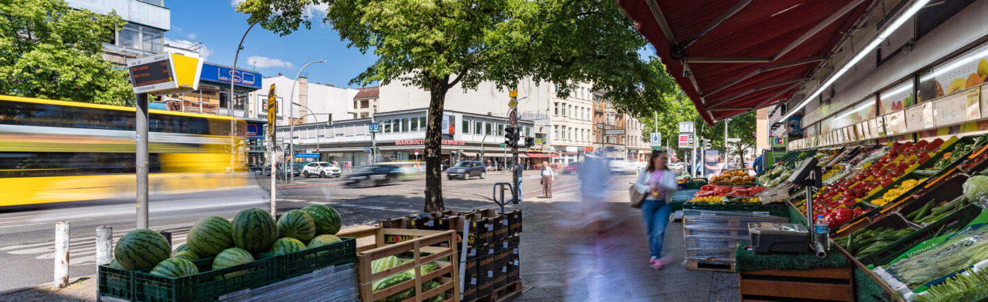 Ein lebhafter Straßenmarkt mit einem frischen Obst- und Gemüsestand auf einem Gehweg. Im Hintergrund ist ein gelber Bus in Bewegung, es sind mehrere Gebäude zu sehen und eine digitale Bushaltestellenanzeige. Ein sonniger Tag mit grünem Baumlaub und blauem Himmel.