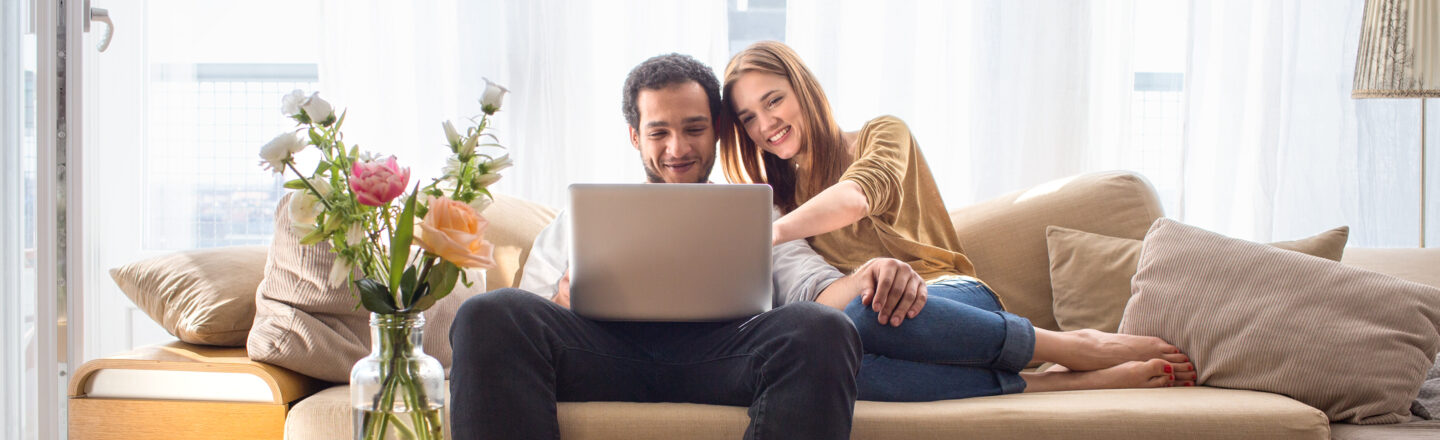 junges Paar sitzt entspannt im Wohnzimmer auf der Couch und schauen sich etwas auf einem Laptop an.