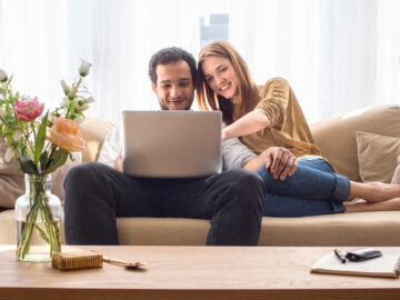 junges Paar sitzt entspannt im Wohnzimmer auf der Couch und schauen sich etwas auf einem Laptop an.