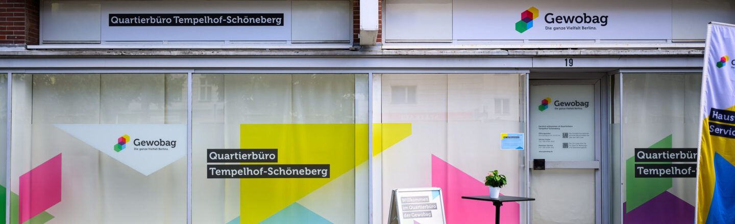 Bunte Schaufensterfront des Gewobag-Quartierbüros Tempelhof-Schöneberg mit Logo, Eingangstür, Infoaufsteller und kleinem Tisch mit Pflanze.