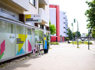 Straßenansicht mit buntem Gewobag-Quartierbüro Tempelhof-Schöneberg, Pflanzkübel und Aufsteller, dahinter Gehweg, Kreuzung und Häuser.