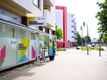 Straßenansicht mit buntem Gewobag-Quartierbüro Tempelhof-Schöneberg, Pflanzkübel und Aufsteller, dahinter Gehweg, Kreuzung und Häuser.