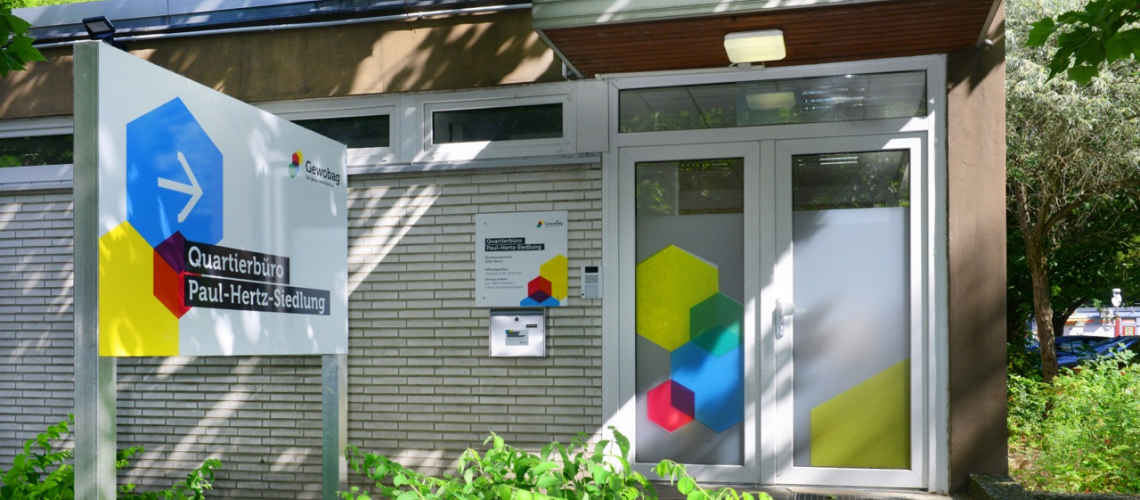 Eingang des Quartierbüros Paul-Hertz-Siedlung mit Schild und Glastür, beide mit bunten geometrischen Formen gestaltet.