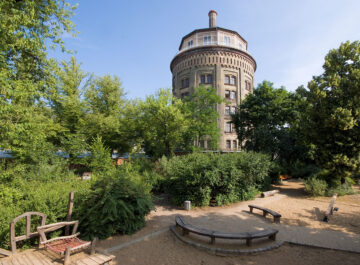 Runder Backsteinturm ragt über grünem Park mit Sandspielplatz, Bänken und Holzspielgeräten in den blauen Himmel.