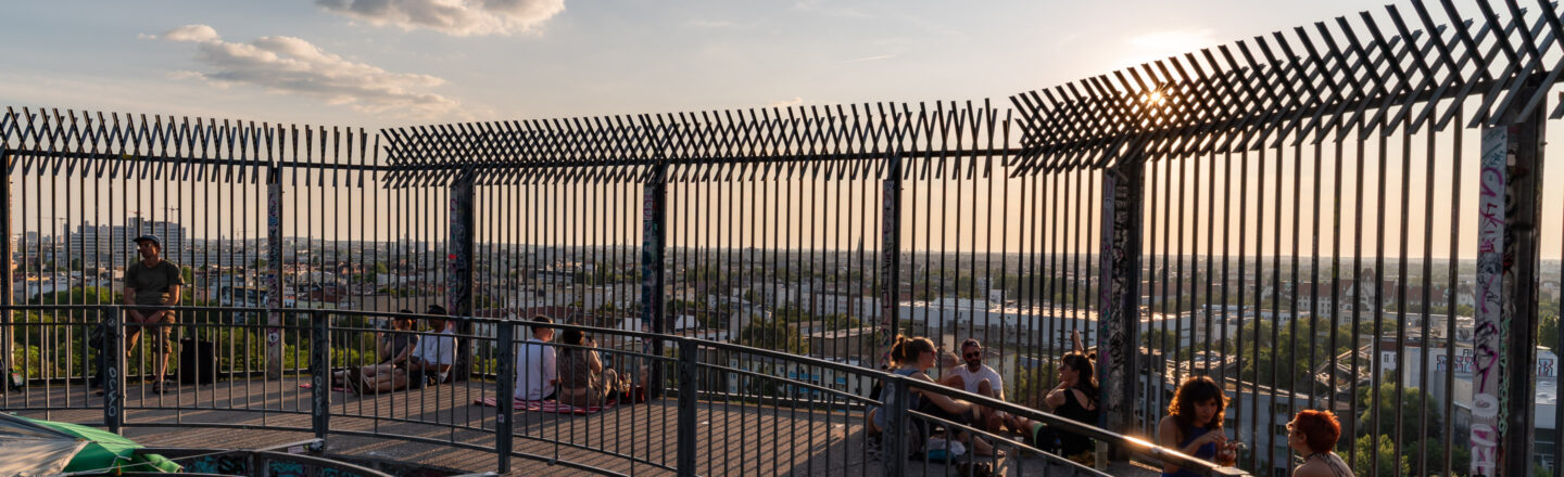 Aussichtsplattform mit hohen Gitterzäunen bei Sonnenuntergang, mehrere Menschen sitzen und entspannen, Stadtlandschaft im Hintergrund.