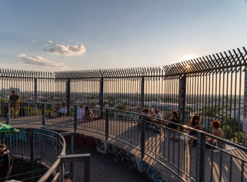 Aussichtsplattform mit hohen Gitterzäunen bei Sonnenuntergang, mehrere Menschen sitzen und entspannen, Stadtlandschaft im Hintergrund.