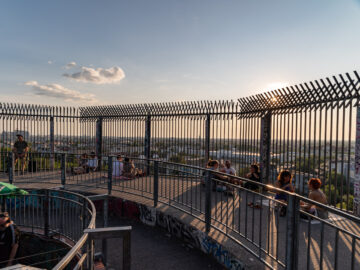 Aussichtsplattform mit hohen Gitterzäunen bei Sonnenuntergang, mehrere Menschen sitzen und entspannen, Stadtlandschaft im Hintergrund.