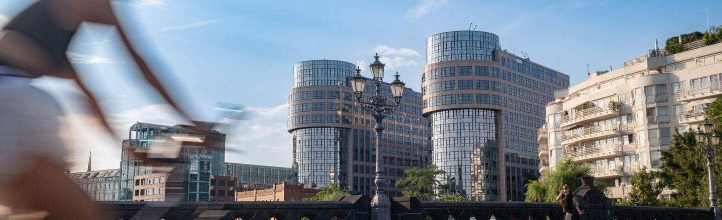 Verschwommener Radfahrer auf Brücke vor modernen Glas- und Bürogebäuden unter blauem Himmel, Fußgängerin rechts, urbaner Stadtblick.