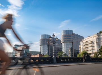 Verschwommener Radfahrer überquert Brücke vor modernen Glasbauten, blauer Himmel und sommerliches Licht prägen die urbane Szene.