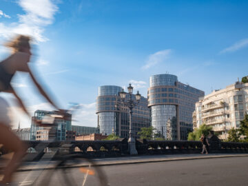 Verschwommener Radfahrer überquert Brücke vor modernen Glasbauten, blauer Himmel und sommerliches Licht prägen die urbane Szene.