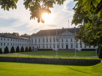 Schloss Bellevue im Abendlicht: weiße Fassade, weiter grüner Rasen mit Fahnenmasten, eingerahmt von Bäumen unter hellem Himmel.