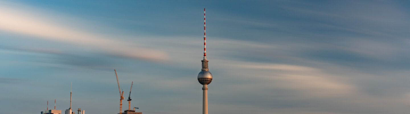 Berliner Fernsehturm ragt vor weitem Himmel mit ziehenden Wolken in die Höhe, unten angedeutete Stadt mit Baukränen.