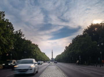 Berliner Straße im Tiergarten mit Autos, gesäumt von dichtem Grün; am Horizont steht die Siegessäule unter bewölktem Himmel.