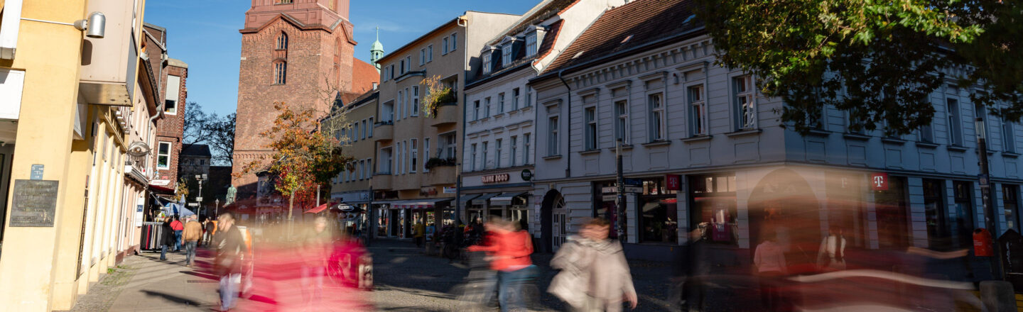 Belebte Altstadtstraße mit historischen Wohnhäusern und rotem Kirchturm, viele Fußgänger als Bewegungsunschärfe im Sonnenschein.