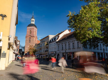 Belebter Platz in der Altstadt mit rotem Kirchturm, historischen Fassaden und vielen verschwommenen Passanten im sonnigen Herbstlicht.