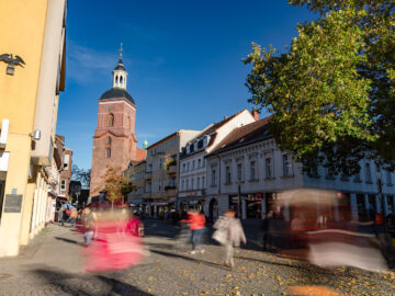 Belebter Platz in der Altstadt mit rotem Kirchturm, historischen Fassaden und vielen verschwommenen Passanten im sonnigen Herbstlicht.