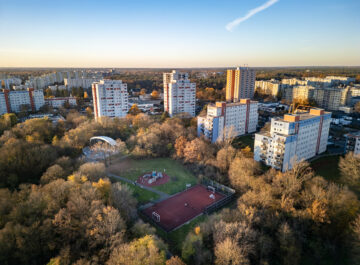 Luftaufnahme: mehrgeschossige Wohnhäuser umgeben von herbstlichen Bäumen, unten Sportplatz und Spielbereich in einem grünen Quartier.