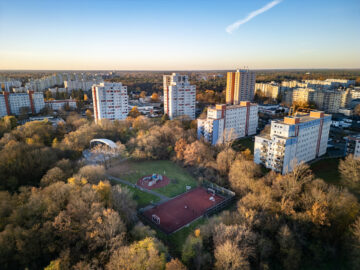 Luftaufnahme: mehrgeschossige Wohnhäuser umgeben von herbstlichen Bäumen, unten Sportplatz und Spielbereich in einem grünen Quartier.