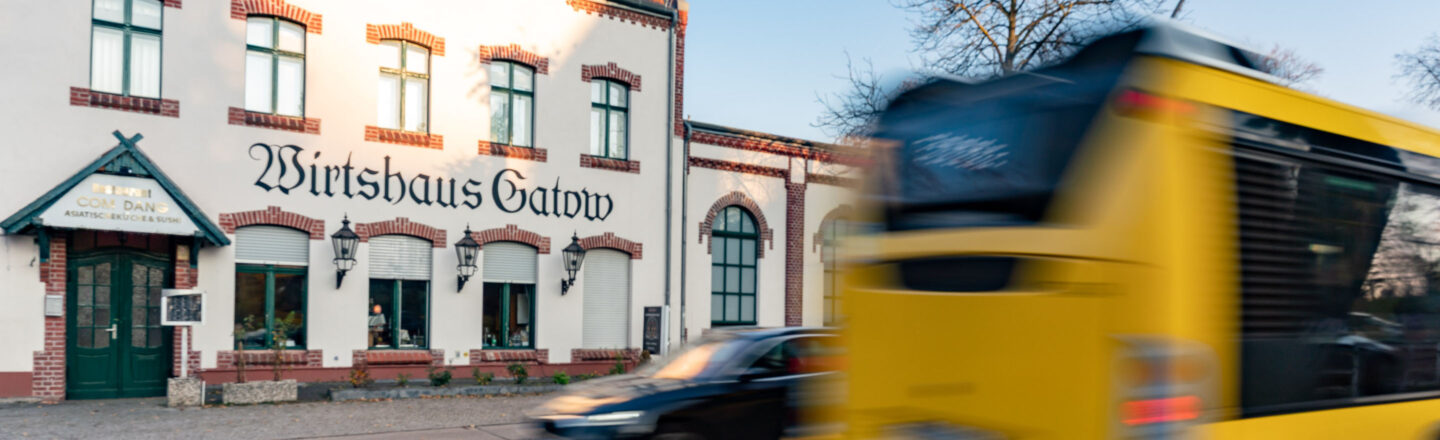 Vor dem historischen Gebäude „Wirtshaus Gatow“ fährt ein gelber Linienbus vorbei, daneben stehen parkende Autos an der Straße.