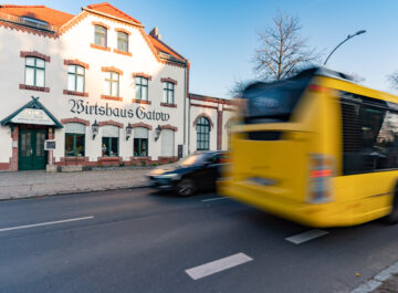 Vor dem historischen Gebäude „Wirtshaus Gatow“ fährt ein gelber Linienbus vorbei, ein Auto daneben auf der Straße, Bäume im Hintergrund.
