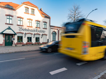 Vor dem historischen Gebäude „Wirtshaus Gatow“ fährt ein gelber Linienbus vorbei, ein Auto daneben auf der Straße, Bäume im Hintergrund.