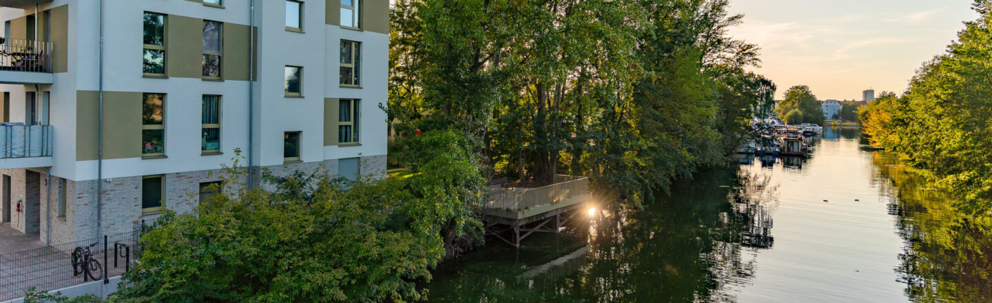 Mehrfamilienhaus mit Balkonen direkt am Fluss, daneben grüne Bäume, Steg über dem Wasser und Bootsanleger im Abendlicht.