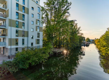 Mehrfamilienhaus mit Balkonen direkt am Fluss, daneben grüne Bäume, Steg über dem Wasser und Bootsanleger im Abendlicht.