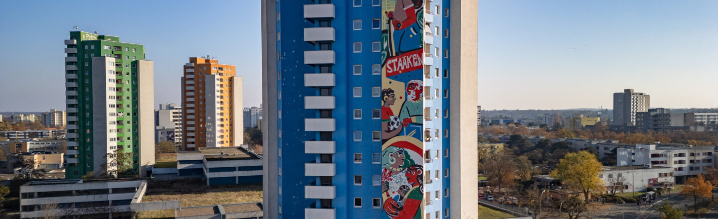 Bunte Hochhäuser, im Vordergrund blaues Wohnhaus mit großem Wandbild „Staaken“, dahinter weitere Türme und Stadtlandschaft.
