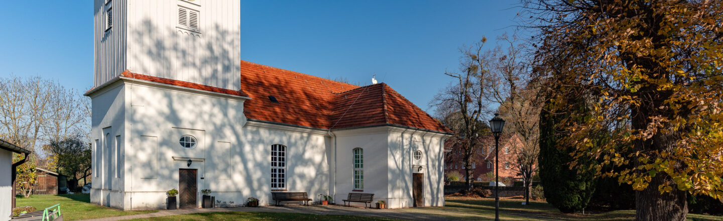 Weiße Dorfkirche mit rotem Ziegeldach, davor Wiese und herbstliche Bäume unter blauem Himmel.