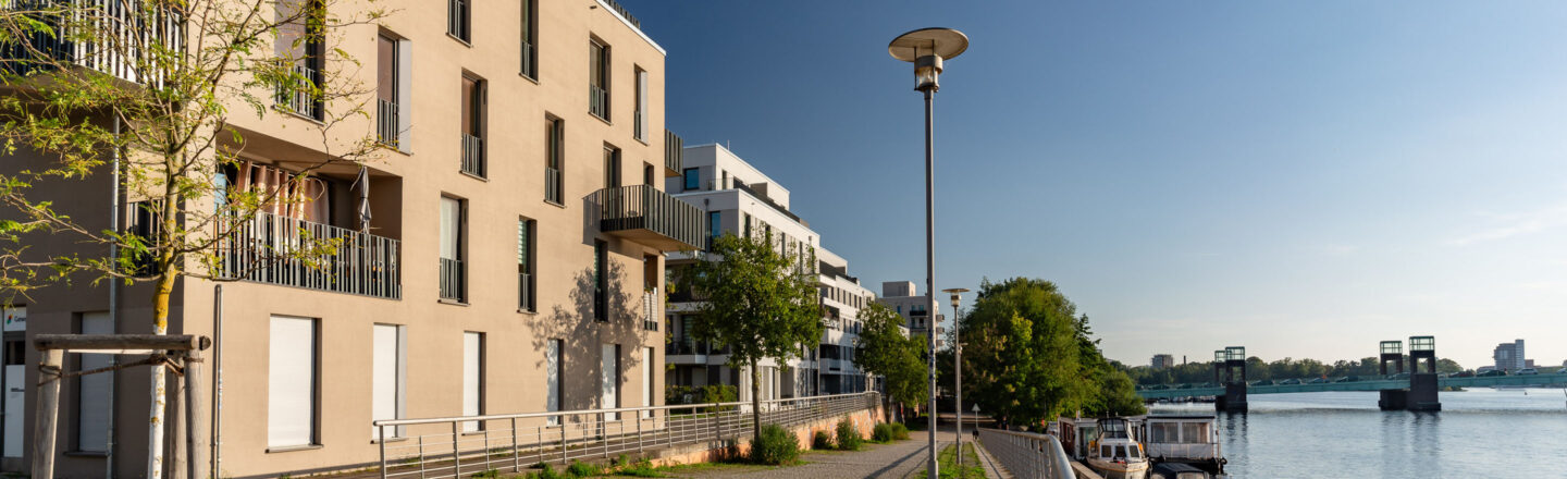 Moderne Wohnhäuser mit Balkonen an einer Uferpromenade, unten Boote am Steg, rechts eine Brücke über den ruhigen Fluss.
