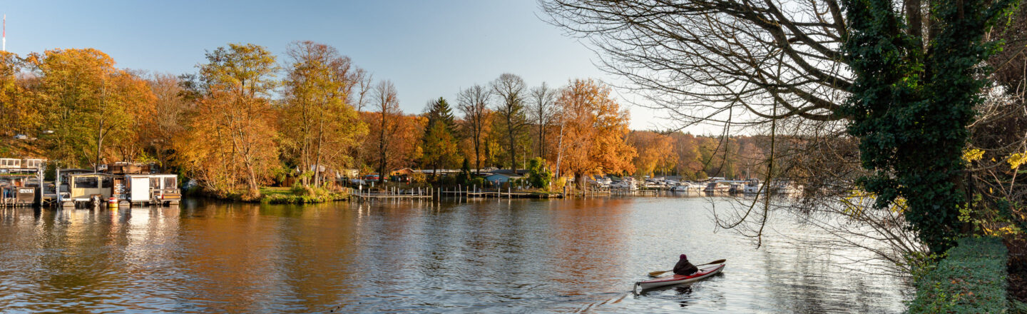 Herbstliche Flusslandschaft mit bunten Bäumen, Bootsstegen und vielen Booten; vorne paddelt eine Person im Kajak über das ruhige Wasser.