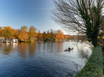 Herbstliche Flusslandschaft mit bunten Bäumen und Bootsstegen; rechts fährt eine Person im Kajak über das ruhige Wasser.
