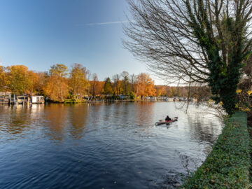 Herbstliche Flusslandschaft mit bunten Bäumen und Bootsstegen; rechts fährt eine Person im Kajak über das ruhige Wasser.