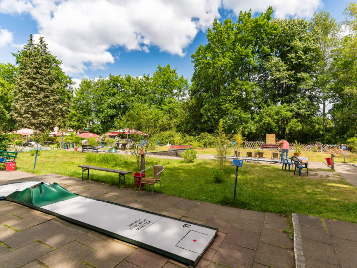 Outdoor-Minigolfplatz bei sonnigem Wetter mit grüner Wiese und großen Bäumen im Hintergrund. Menschen spielen und genießen die Umgebung, die von Sonnenschirmen und Gartenmöbeln ergänzt wird. Ein plattierter Weg führt durch die Anlage.