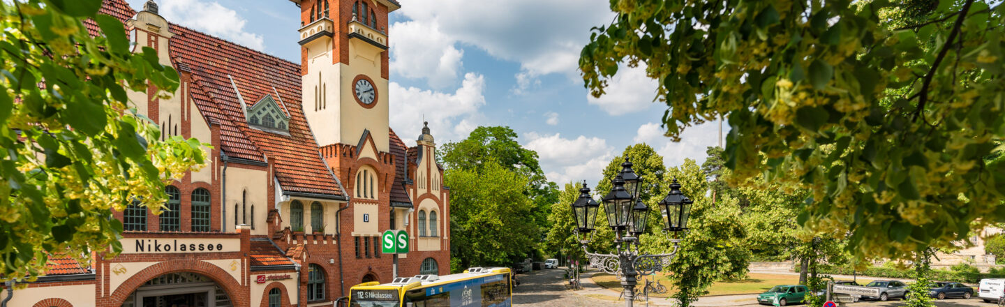 Das Bild zeigt den Bahnhof Berlin-Nikolassee mit seinem markanten roten Ziegeldach und einem Uhrenturm. Vor dem Gebäude steht ein Bus, und das grüne "S" Signet der S-Bahn ist sichtbar. Umgeben ist der Bahnhof von grünen Bäumen und einem dekorativen Straßenlaternenpfosten. Der Himmel ist teils bewölkt.