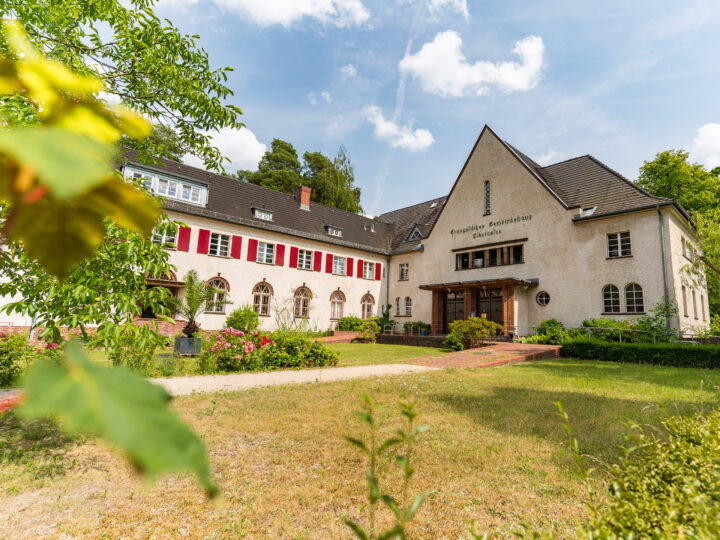 Das Bild zeigt das Evangelische Gemeindehaus Nikolassee, ein zweistöckiges Gebäude mit hellen Fassaden und einem dunklen Dach. Auf der rechten Seite des Hauses steht der Schriftzug "Evangelisches Gemeindehaus Nikolassee". Die Fenster sind teilweise mit roten Fensterläden ausgestattet. Im Vordergrund befindet sich ein gepflegter Garten mit grüner Vegetation und blühenden Blumen. Der Himmel ist leicht bewölkt, und die Sonne scheint hell.