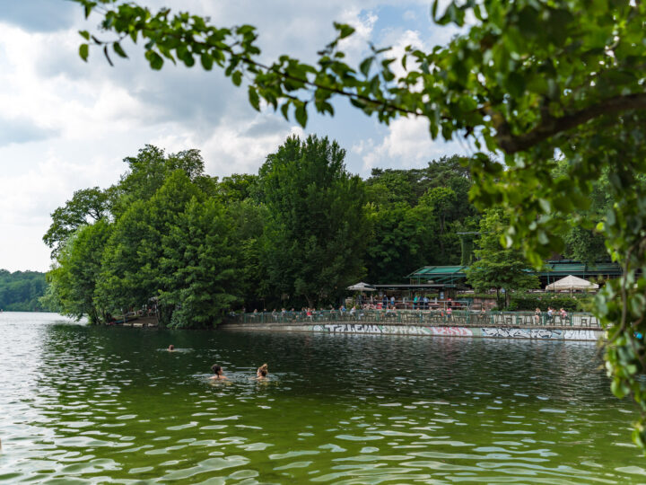 Das Bild zeigt einen See mit mehreren Menschen, die im Wasser schwimmen. Im Hintergrund ist eine Uferpromenade mit graffiti-bedeckter Mauer und einer Aussichtsplattform zu sehen, auf der weitere Menschen sitzen und die Umgebung genießen. Der See ist umgeben von dichtem, grünem Wald. Der Himmel ist bewölkt, aber dennoch scheint etwas Sonne durch die Wolken. Einige Baumzweige hängen von oben in das Bild hinein.