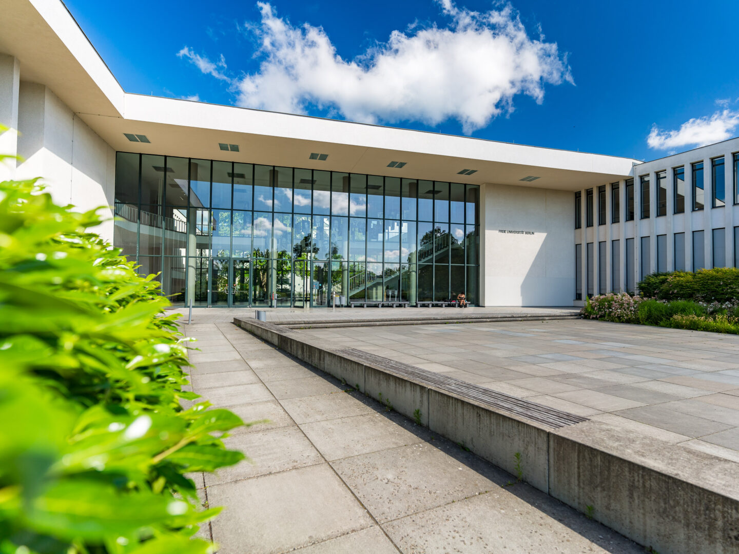 Das Bild zeigt den modernen Eingangsbereich eines Gebäudes der Freien Universität Berlin. Die Fassade aus Glas und hellem Beton erstreckt sich über zwei Stockwerke und reflektiert den blauen Himmel mit wenigen weißen Wolken. Im Vordergrund sind große quadratische Steinplatten und grüne Sträucher zu sehen. Links sitzen zwei Personen auf dem Boden nahe dem Eingang.