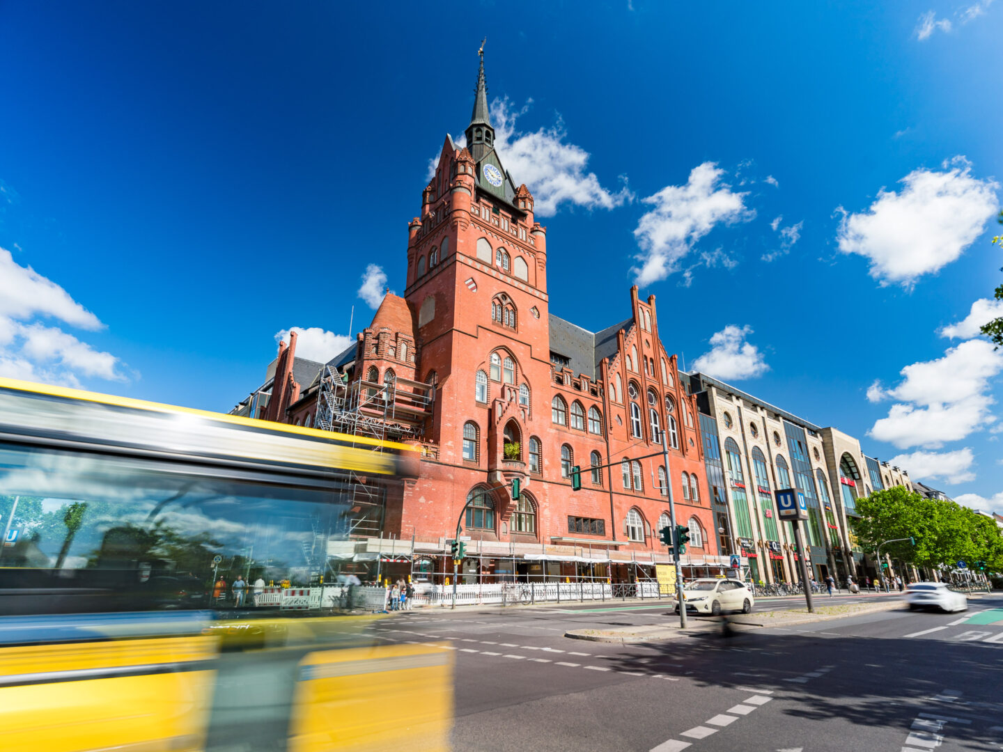 Das Bild zeigt ein ikonisches rotes Backsteingebäude in Berlin, das mit einem steilen Turm und einer Uhr ausgestattet ist. Die Architektur ist historisch und beeindruckend. Im Vordergrund fährt ein gelber Bus vorbei, der das Bild teilweise verwischt. Die Straße ist belebt mit Autos und Fußgängern. Darüber spannt sich ein strahlend blauer Himmel mit vereinzelten weißen Wolken. Rechts im Bild sind weitere Geschäftsgebäude sichtbar, neben einem Eingang zur U-Bahn-Station.