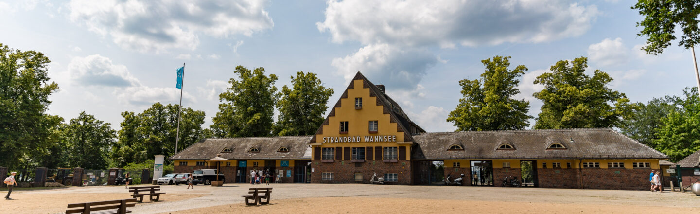 Das Bild zeigt das Eingangsgebäude des Strandbads Wannsee in Berlin. Das Gebäude hat eine gelbe Fassade mit roten Ziegeln und einem Satteldach. Über dem Eingang steht der Schriftzug "Strandbad Wannsee". Im Vordergrund befinden sich Sitzbänke und eine offene Fläche. Einige Personen laufen auf dem Gelände, und Bäume säumen den Hintergrund. Der Himmel ist teilweise bewölkt.