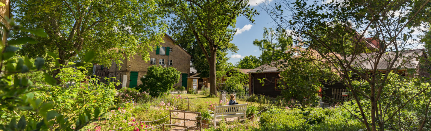 Das Bild zeigt einen idyllischen grünen Garten mit Blumen und Büschen, umgeben von Bäumen. Im Hintergrund sind ein großes Backsteingebäude und ein Holzhaus sichtbar. Eine Person sitzt auf einer weißen Bank und genießt die Szenerie. Der Himmel ist blau mit vereinzelten Wolken.