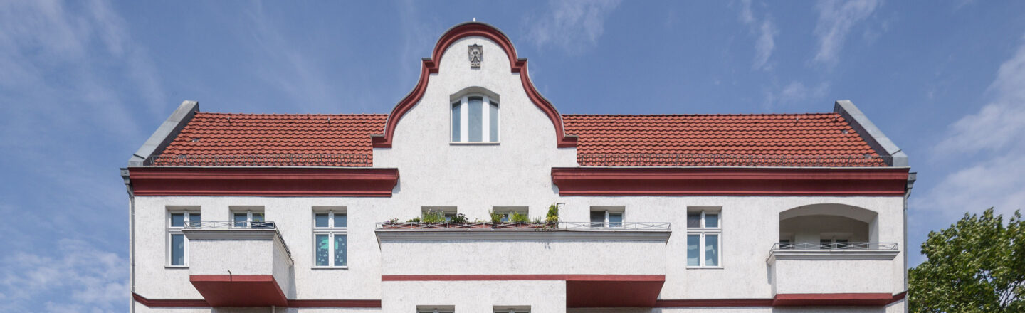 Helle Altbau-Fassade mit rotem Ziegeldach und Balkonen, mittig geschwungener Giebel unter blauem Himmel, etwas Grün auf den Brüstungen.