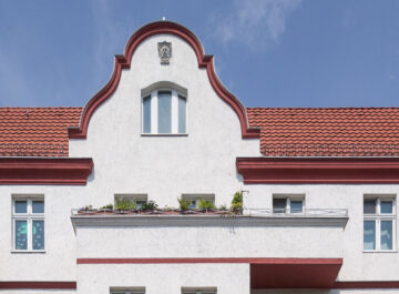 Helle Altbau-Fassade mit rotem Ziegeldach und Balkonen, mittig geschwungener Giebel unter blauem Himmel, etwas Grün auf den Brüstungen.