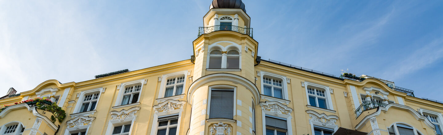 Das Bild zeigt ein mehrstöckiges Gebäude im Jugendstil mit gelber Fassade und kunstvollen Verzierungen. Es besitzt eine markante Turmspitze und mehrere Fenster mit dekorativen Elementen. Einige Bereiche sind mit Pflanzen und Blumen auf den Balkonen geschmückt. Der Himmel ist klar und blau, was dem Gebäude einen lichtdurchfluteten Hintergrund verleiht.