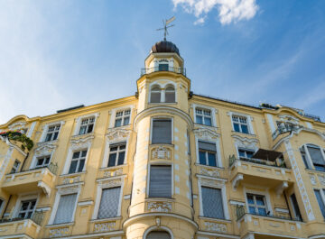 Das Bild zeigt ein mehrstöckiges, gelbes Jugendstilgebäude mit kunstvollen weißen Verzierungen um die Fenster und Balkone. Im Zentrum des Bildes ist ein runder Turm mit einer Kuppel und einer Wetterfahne zu sehen. Der Himmel ist blau und leicht bewölkt. Auf einigen Balkonen sind Pflanzen und Blumen zu erkennen.
