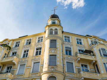 Das Bild zeigt ein mehrstöckiges, gelbes Jugendstilgebäude mit kunstvollen weißen Verzierungen um die Fenster und Balkone. Im Zentrum des Bildes ist ein runder Turm mit einer Kuppel und einer Wetterfahne zu sehen. Der Himmel ist blau und leicht bewölkt. Auf einigen Balkonen sind Pflanzen und Blumen zu erkennen.